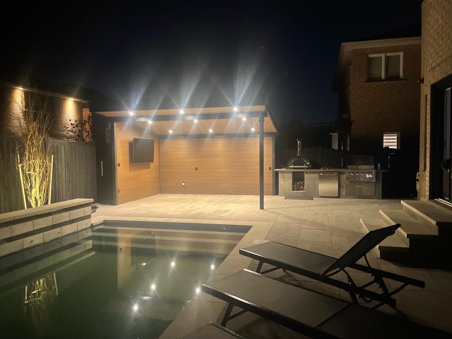 Night view of a backyard with a lit covered patio, outdoor kitchen, pool, and two lounge chairs.