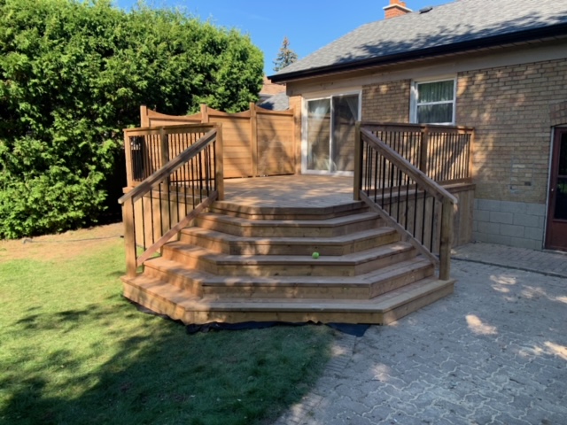 Wooden deck with wide stairs and railings attached to a brick house, surrounded by grass and bushes.