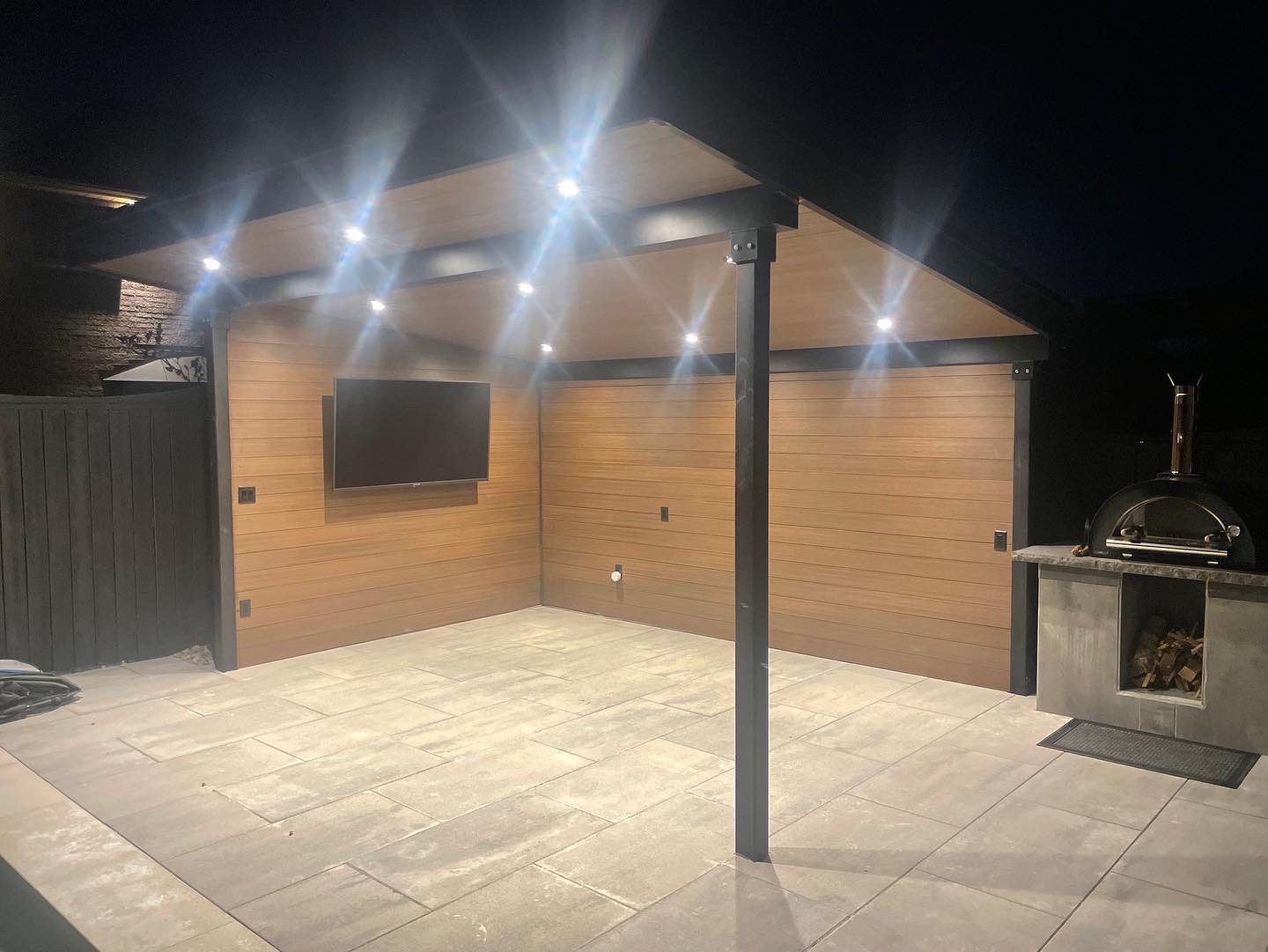 Modern outdoor patio with wood paneling, mounted TV, overhead lights, and pizza oven at night.