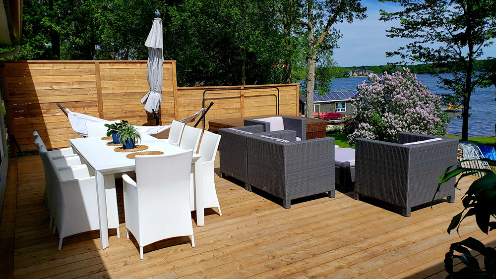 Spacious wooden deck with white dining set, hammock, and gray outdoor lounge furniture overlooking a lakeside garden.