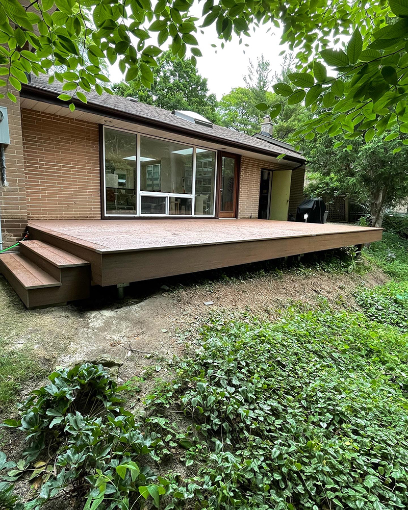 Raised wooden deck with steps attached to a brick house surrounded by greenery and trees.