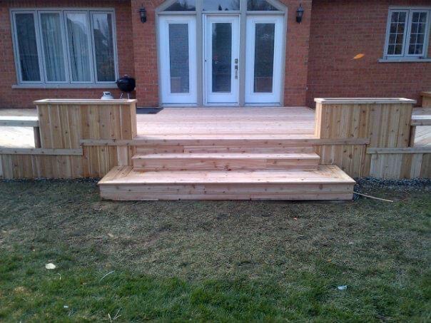 Newly constructed wooden deck with three steps leading to double glass doors on a brick house.