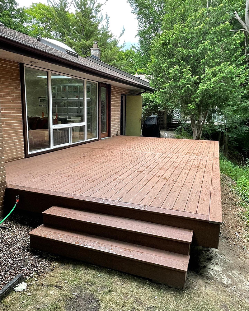 Brown wooden deck with steps attached to a brick house beside green trees and grass.