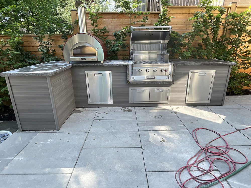 Outdoor kitchen with stainless steel pizza oven, gas grill with open lid, storage cabinets, and gray countertop.