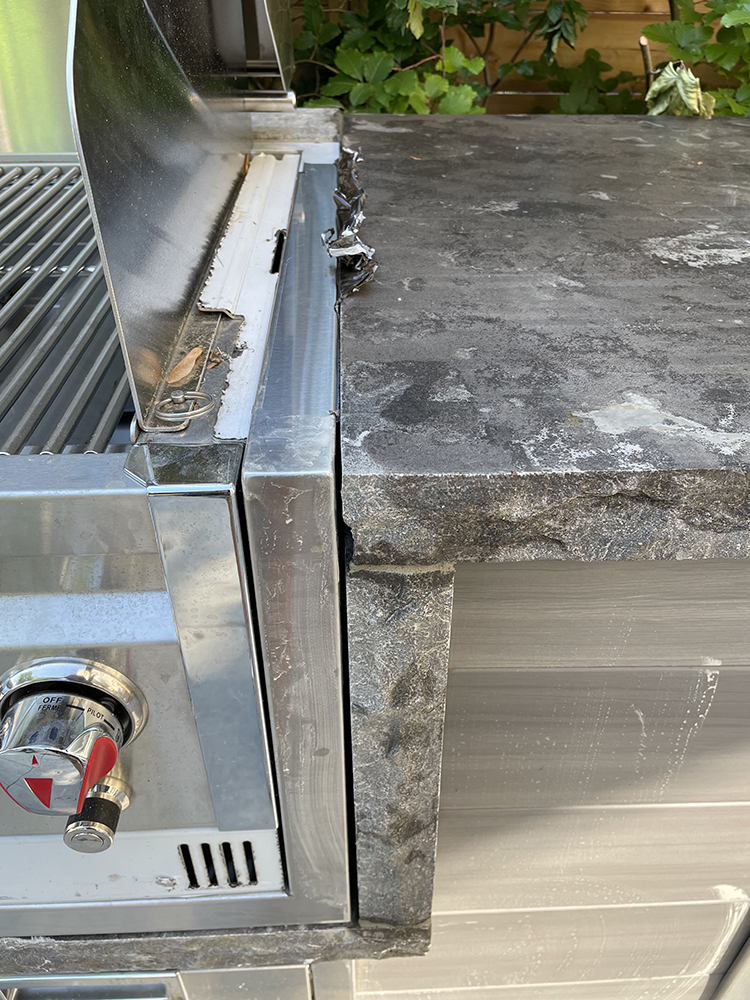 Close-up of a stainless steel gas grill control knob next to a rough textured stone countertop edge.