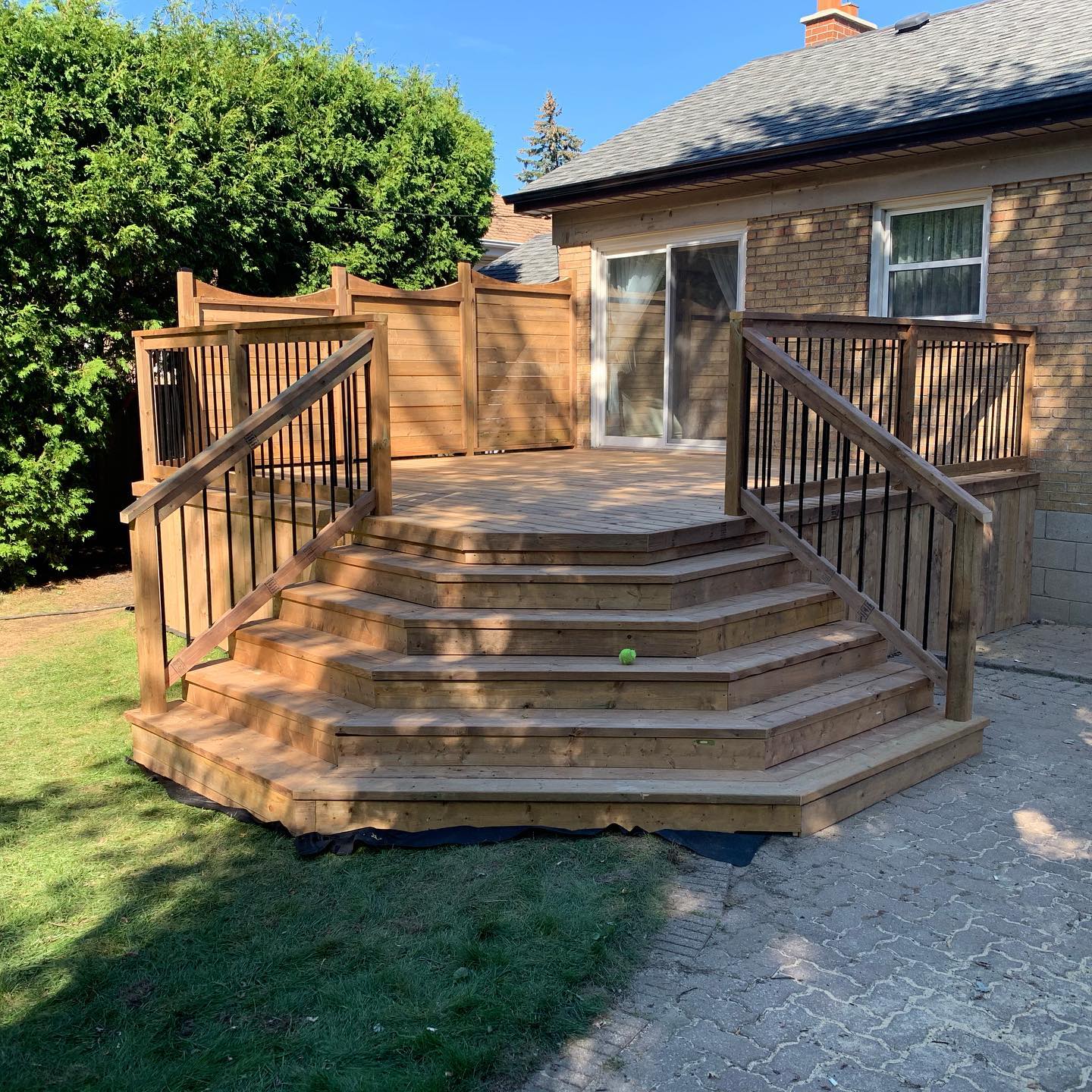 Wooden backyard deck with multi-level hexagonal steps, black metal railings, and privacy fence attached to brick house.