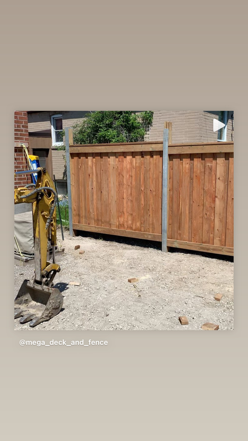Partially constructed wooden fence with metal posts next to a yellow excavator bucket on a dirt ground.