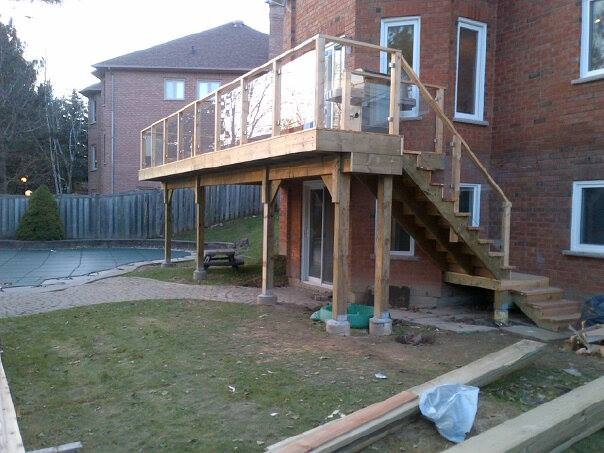 Wooden elevated deck with glass railing and stairs attached to a brick house above a backyard patio.