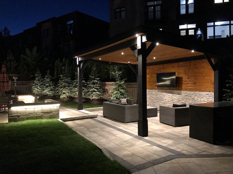 Outdoor covered patio with seating, wall-mounted TV, and soft night lighting in a backyard.