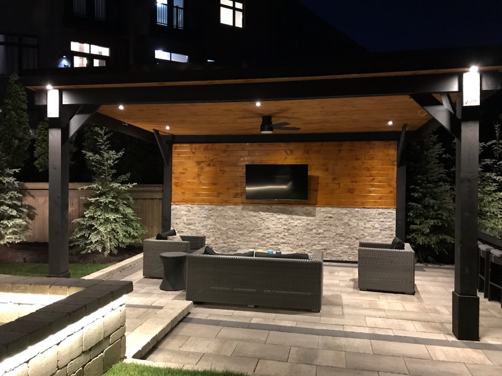 Modern outdoor patio at night with a pergola, cushioned wicker seating, mounted TV, and ambient lighting.