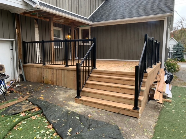 New wooden deck with black metal railing and stairs attached to a house with gray siding.