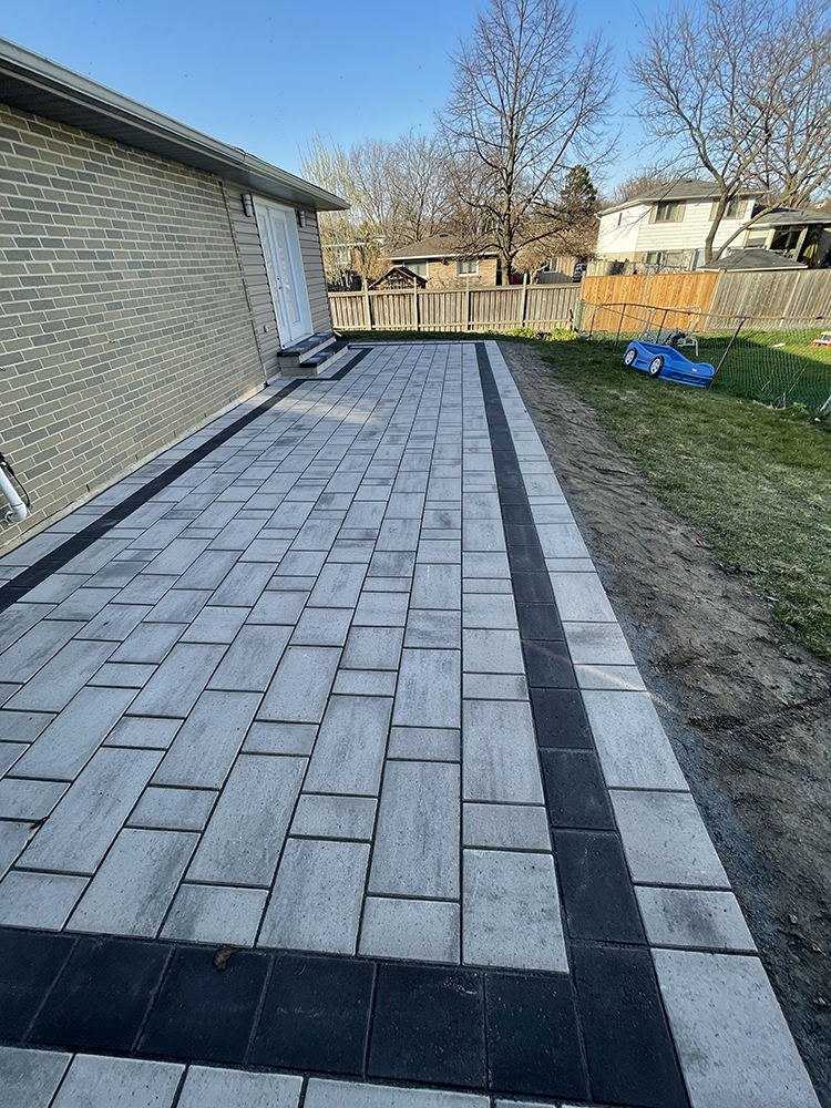Outdoor patio with grey and black rectangular paving stones next to a brick house under a clear blue sky.