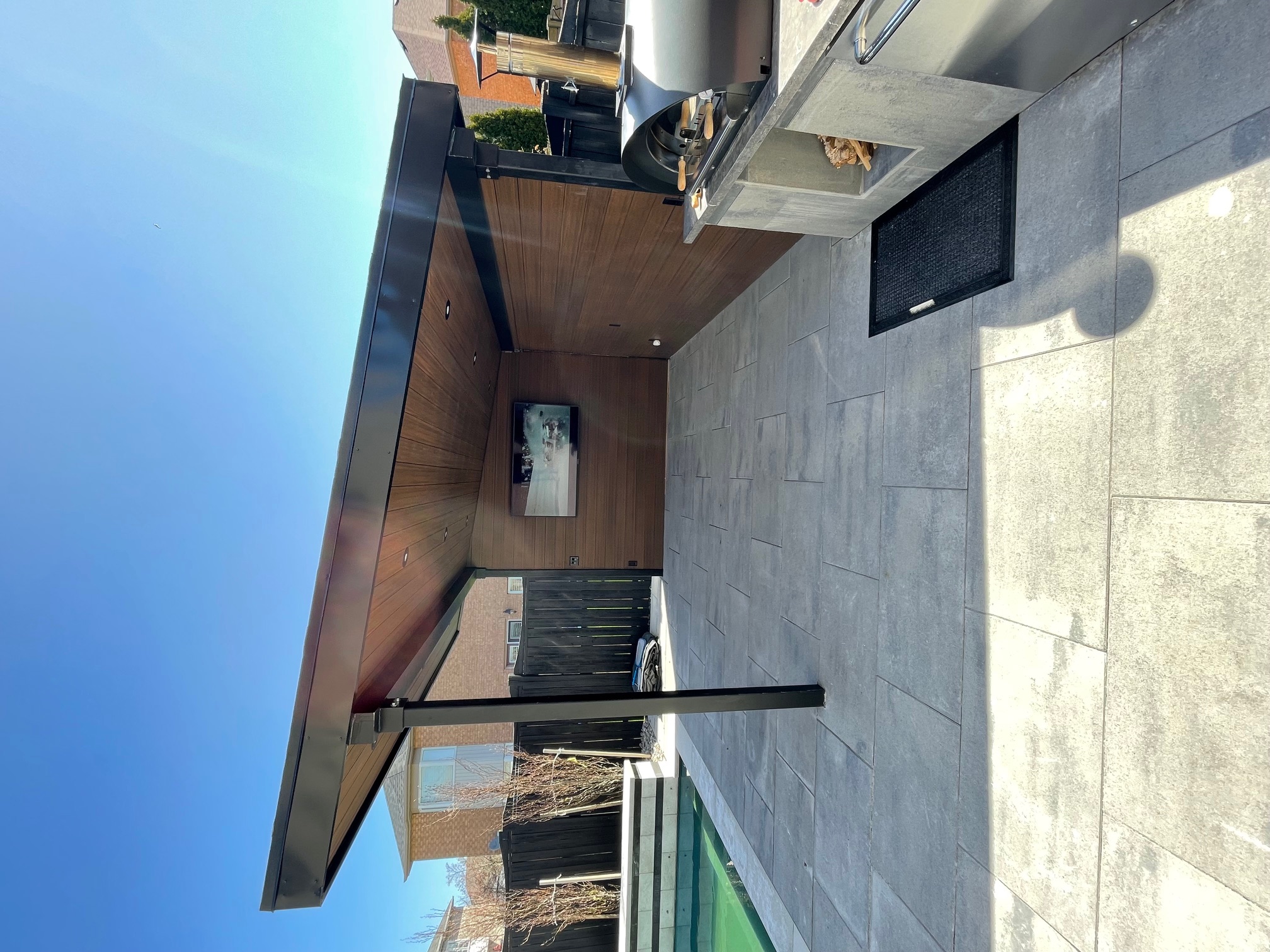 Modern outdoor patio with stone tile flooring, covered seating area with wood paneling, TV mounted, and adjacent pool.