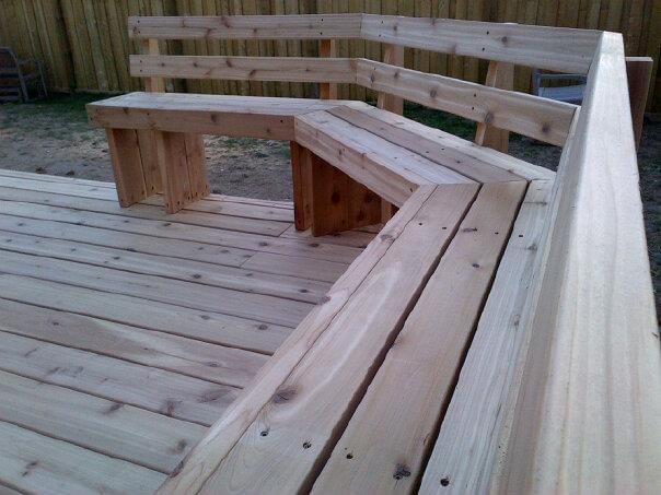 Corner of a wooden deck with attached L-shaped wooden bench seating and backrest.