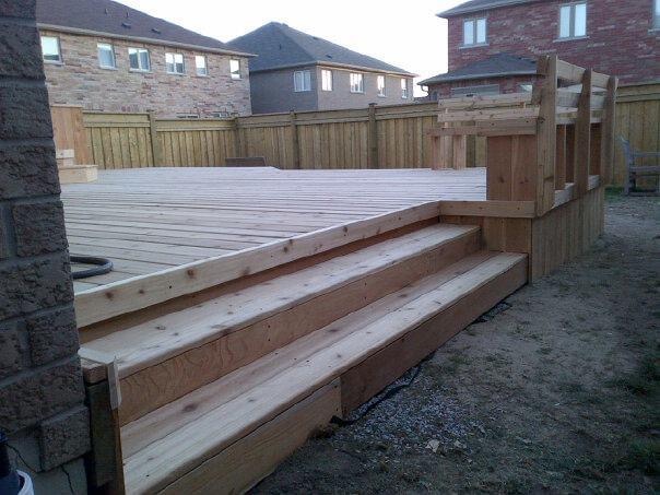 Newly built wooden deck with wide steps and railing in a backyard surrounded by wooden fence and houses.