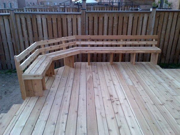 Wooden outdoor deck with built-in L-shaped bench seating against a wooden fence.
