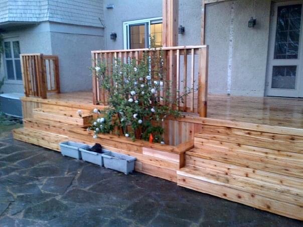 Wooden deck attached to a house with built-in planter holding flowering plants on stone patio.