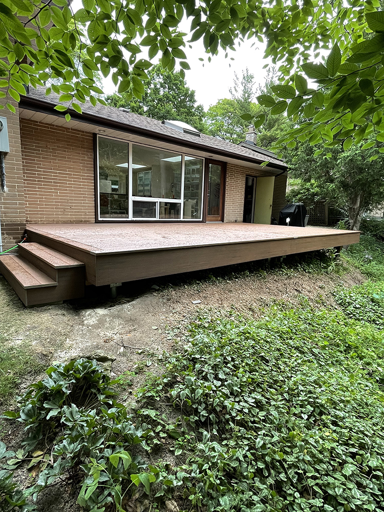 Elevated wooden deck with steps attached to the back of a brick house surrounded by green foliage.