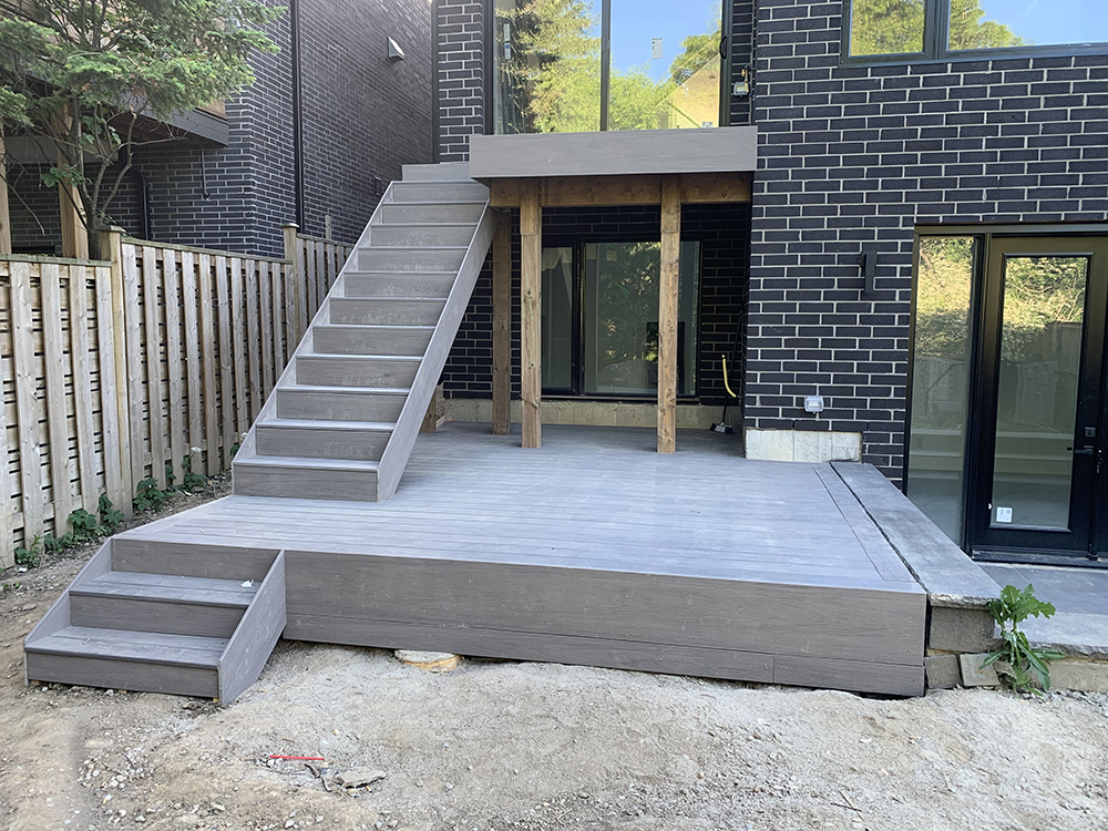 Modern wooden deck with stairs leading to a second level against a dark brick house exterior.