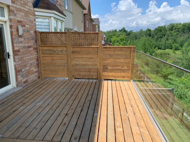 Wooden deck with new and old wood sections, wooden privacy fencing on one side and glass railing overlooking green trees.