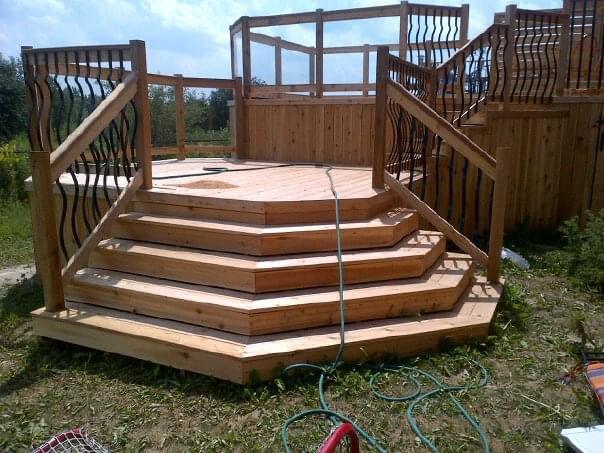 Multi-level wooden deck with hexagonal steps and black iron railings in an outdoor grassy area.