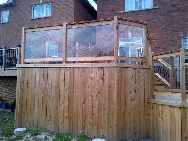 Wooden deck with glass railings attached to a brick house, elevated over a yard.