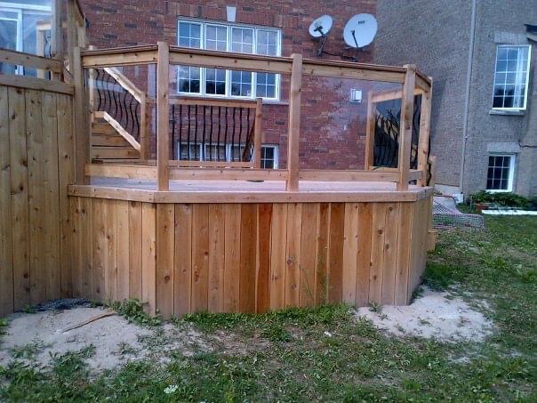 A wooden deck with vertical plank sides and railings with black metal balusters, set against a brick and stucco house.