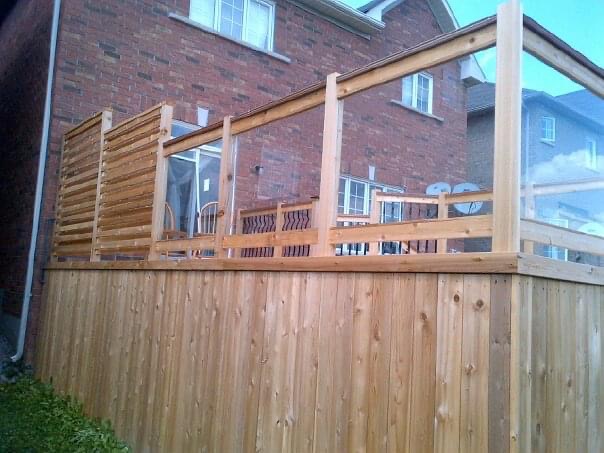 Wooden deck with transparent glass panels and privacy lattice attached to a red brick house.