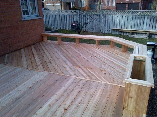Wide wooden deck with built-in seating and planter adjacent to a brick house and fenced backyard.