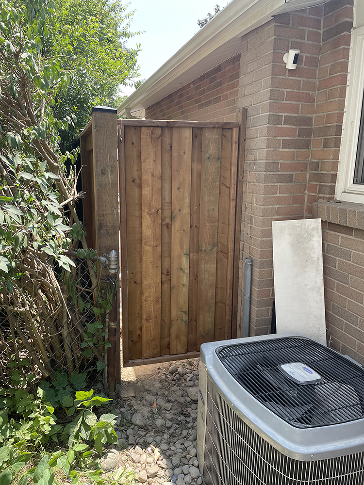 Outdoor wooden gate open beside a brick house wall with an air conditioning unit and green foliage.