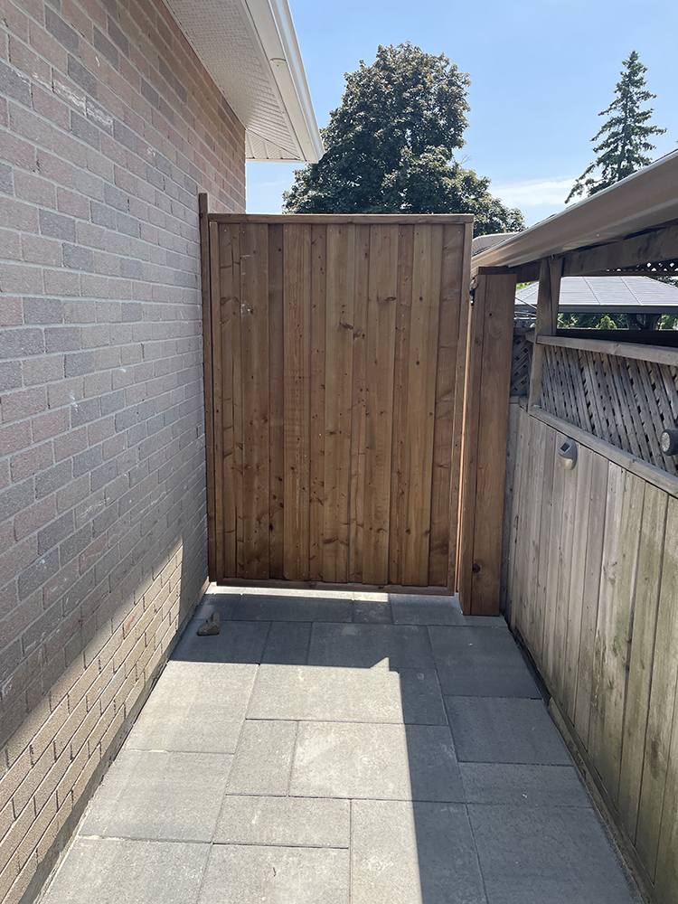 Closed wooden gate between a brick house wall and a wooden fence under a clear blue sky.