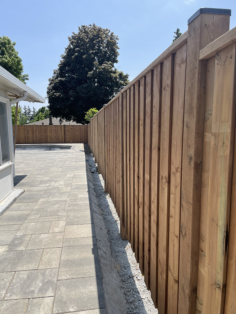 Long wooden fence alongside a paved patio next to a house under a clear blue sky.