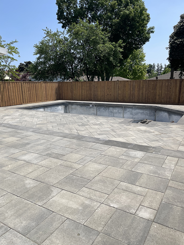 Empty in-ground pool surrounded by a spacious stone patio with a wooden fence and leafy trees in the background.
