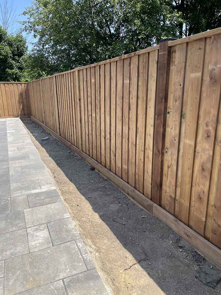 New wooden fence along a bare dirt strip next to a gray paved patio under a clear sky.