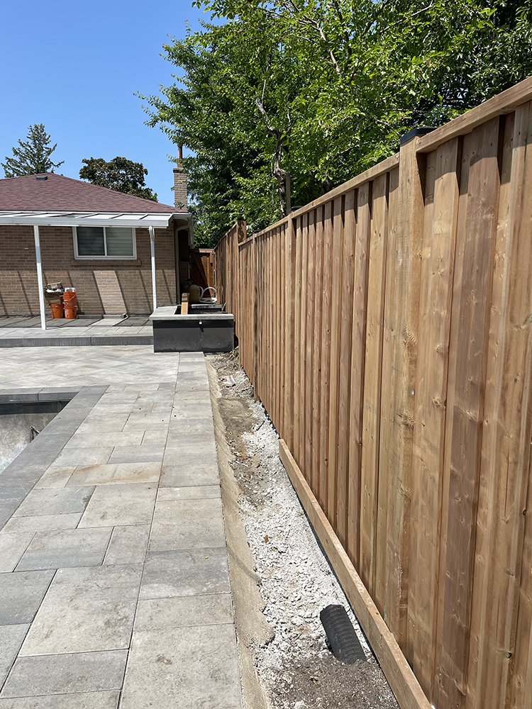 New wooden fence alongside a stone-paved backyard with garden preparation and house in background.
