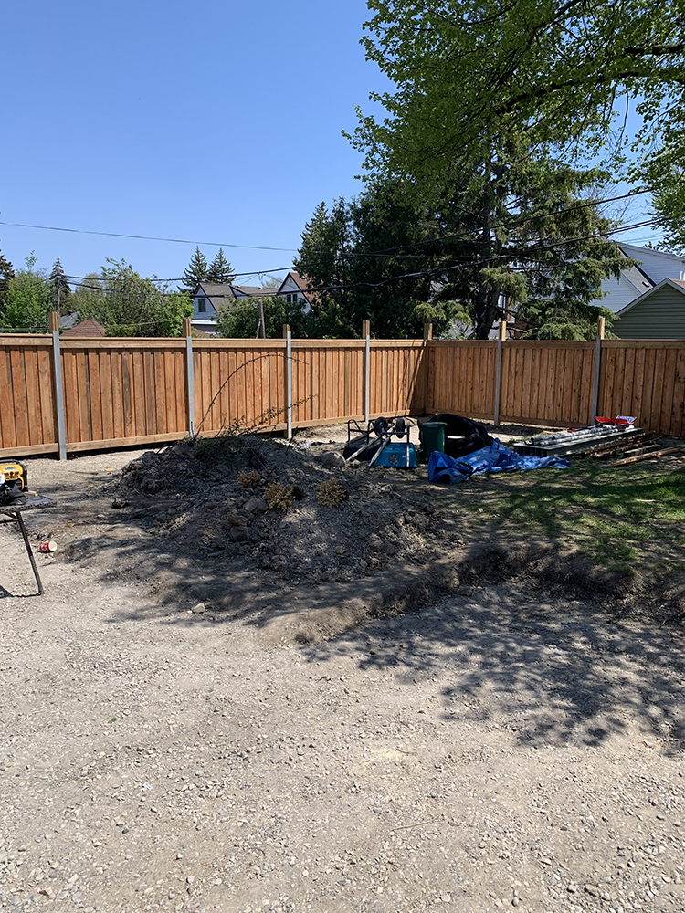 Backyard with new wooden fence, dirt piles, tools, and scattered construction materials under clear blue sky.