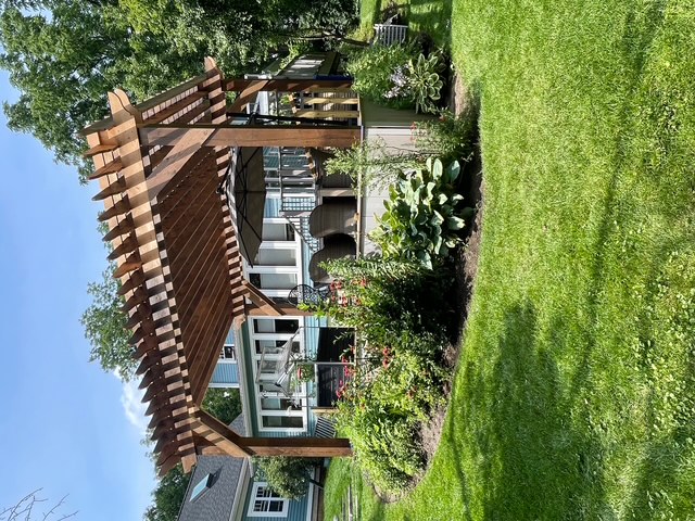 Wooden pergola over a backyard deck with patio furniture and garden plants surrounding it on a sunny day.