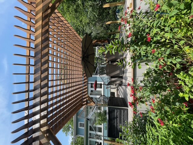 Wooden pergola over a patio with outdoor chairs and umbrella, surrounded by green plants and red flowers.