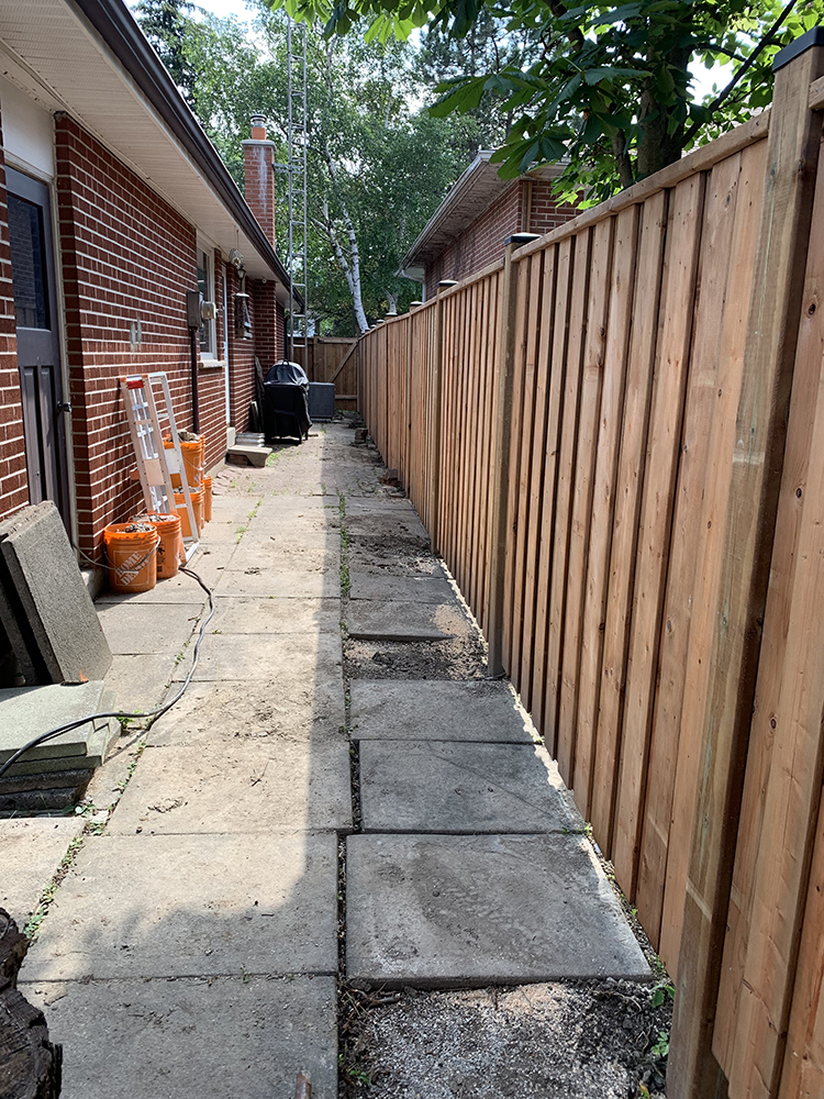 Narrow backyard walkway with concrete paving stones, wooden fence on right, and brick house with tools on left.