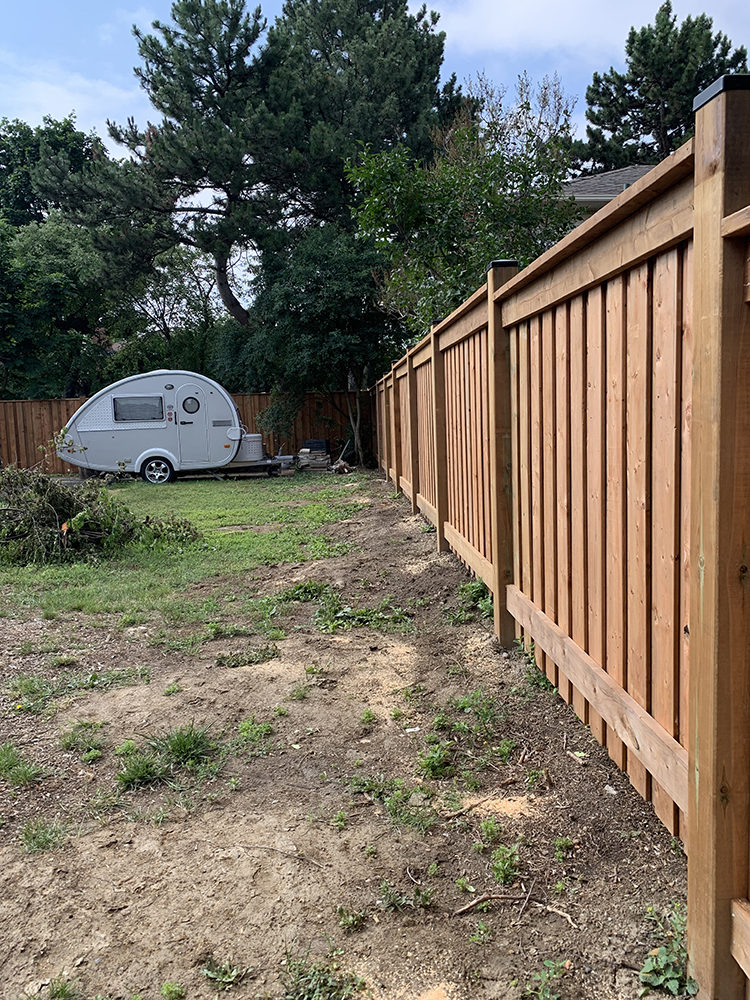 Small white teardrop camper in a backyard with wooden fence and tall trees.