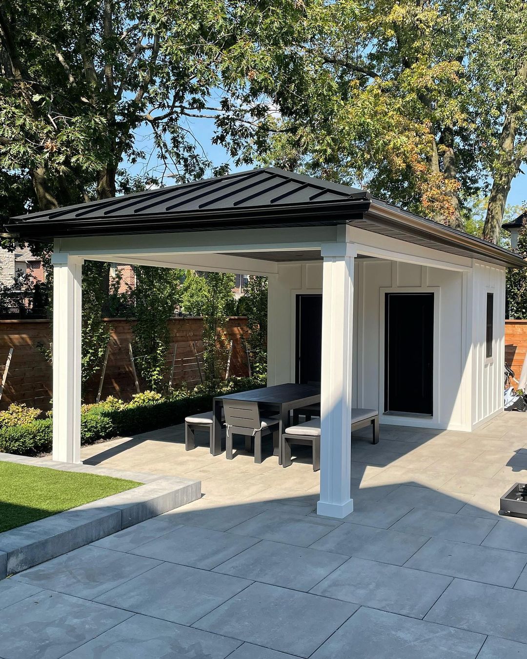 Modern outdoor gazebo with black metal roof and white pillars, featuring a dining table and benches on a stone patio.