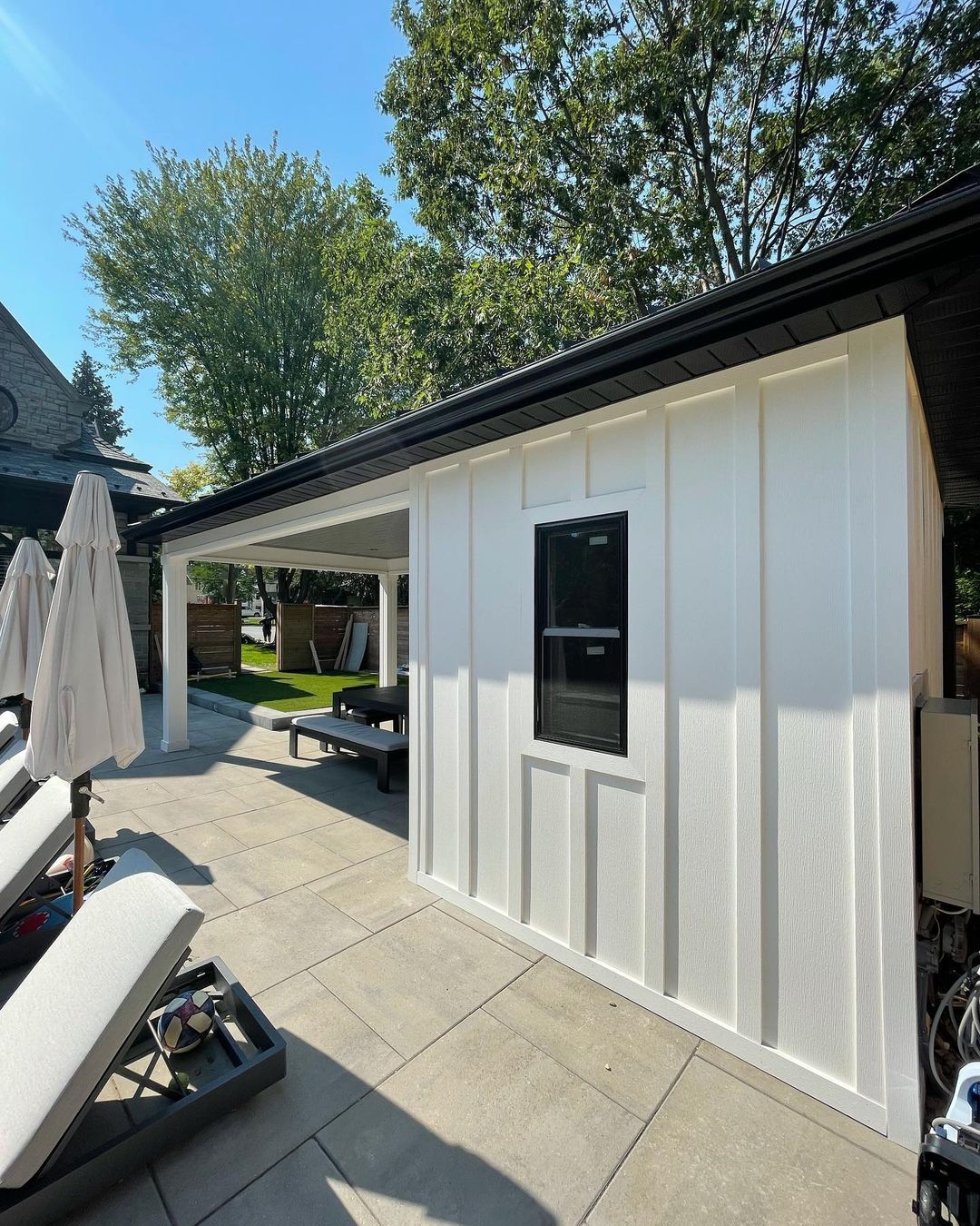 Modern white garden shed with black window and patio seating area under an extended roof on a sunny day.