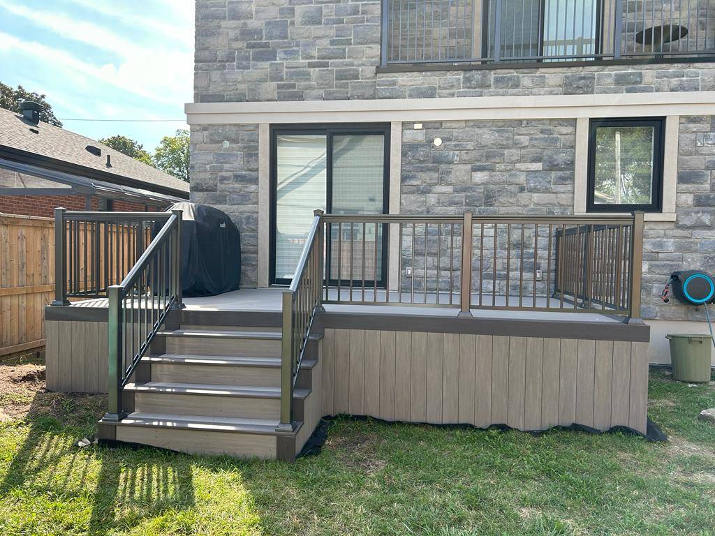 Small elevated backyard deck with stairs, black metal railing, and a covered grill against a stone house wall.